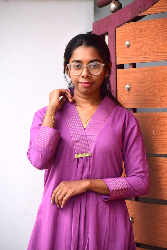Woman wearing a purple traditional outfit with glasses, standing against a wooden panel background.