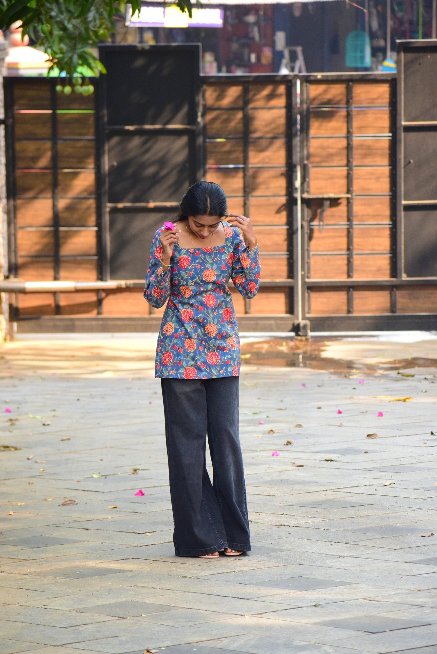 Woman in a colorful floral top and dark pants standing on a paved area with a metal gate in the background.