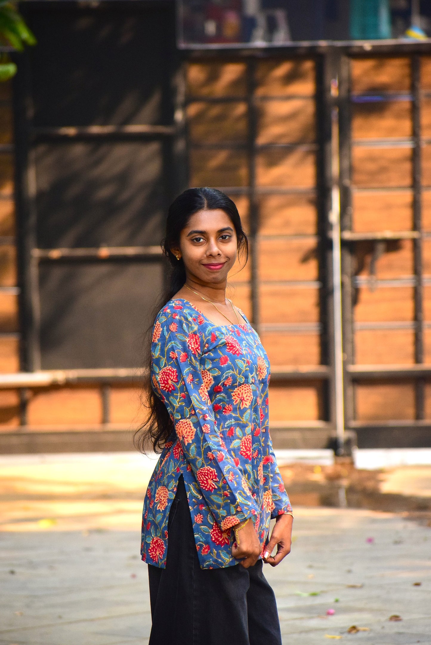 Woman wearing a colorful floral blouse standing outdoors with a wooden structure in the background