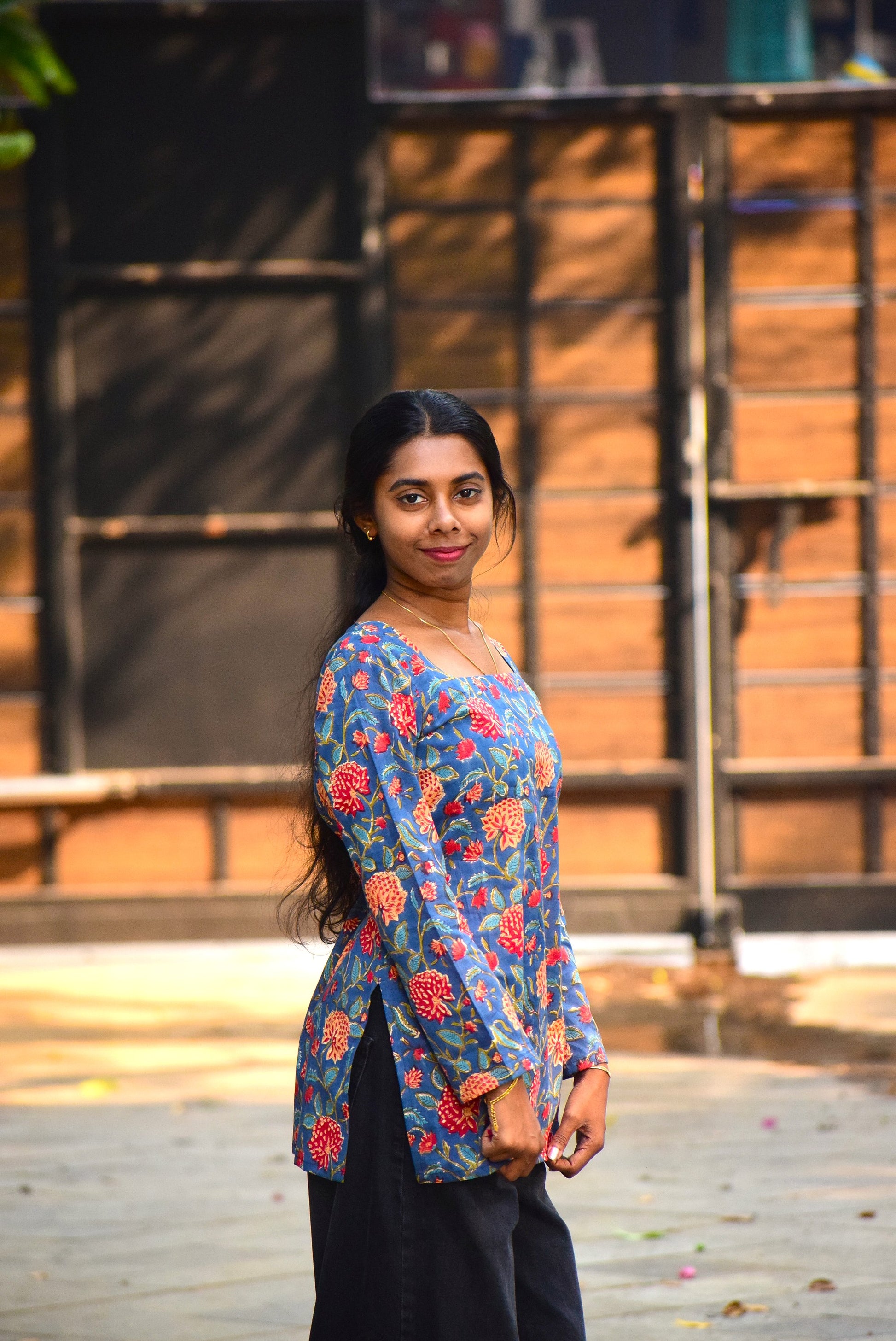Woman wearing a colorful floral blouse standing outdoors with a wooden structure in the background