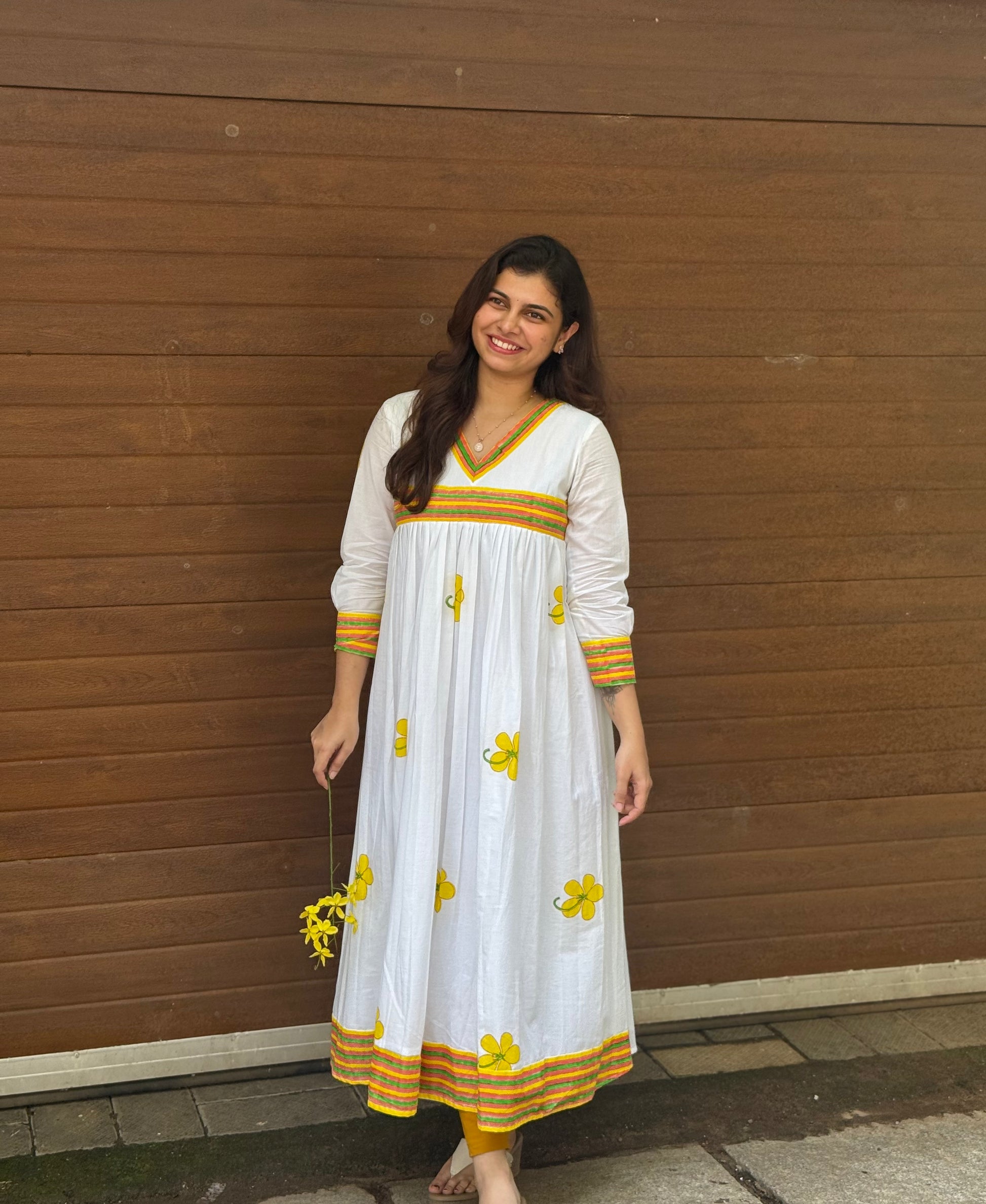 Woman wearing a white dress with yellow floral patterns against a wooden wall.
