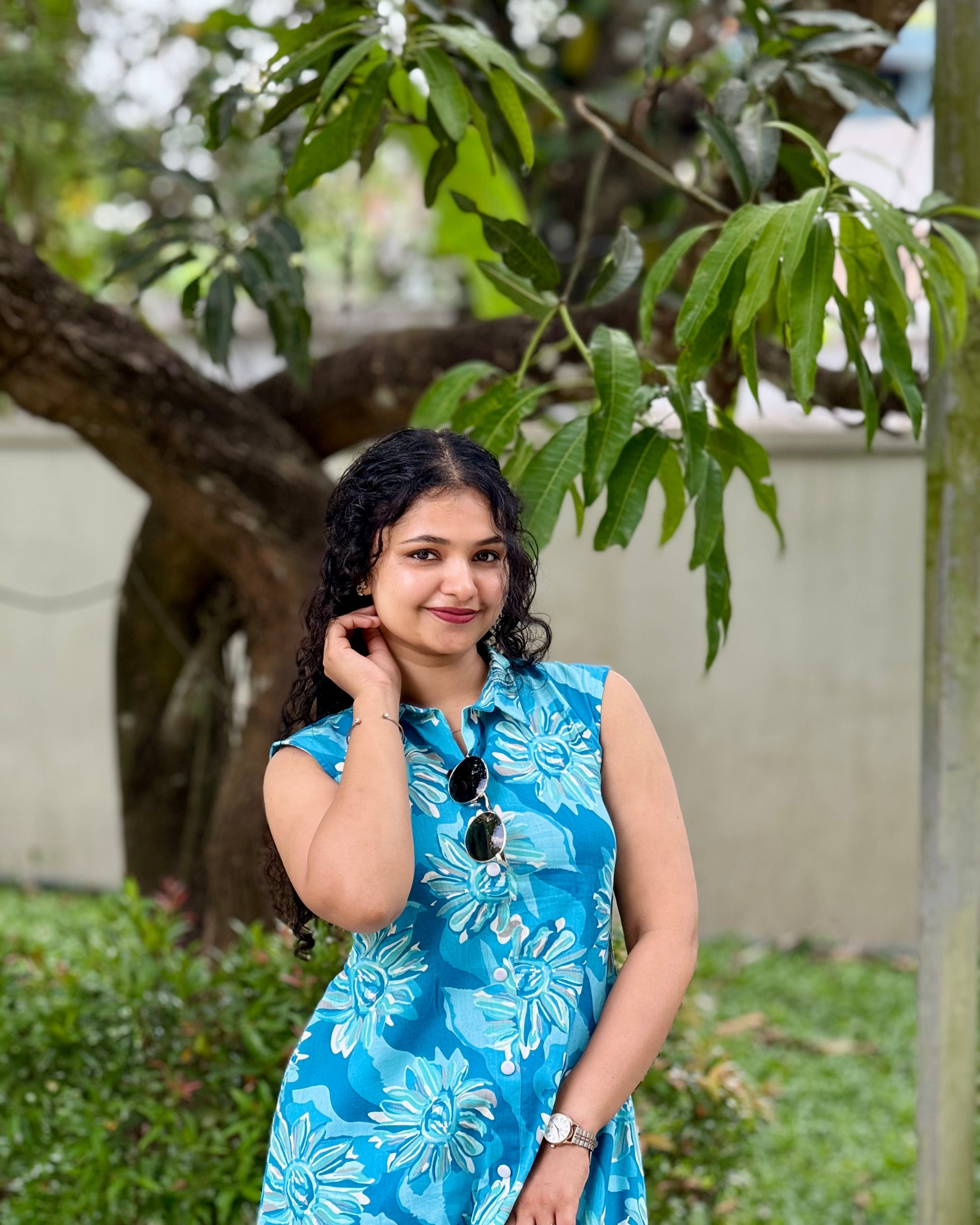 Woman in a blue floral dress standing outdoors with greenery in the background