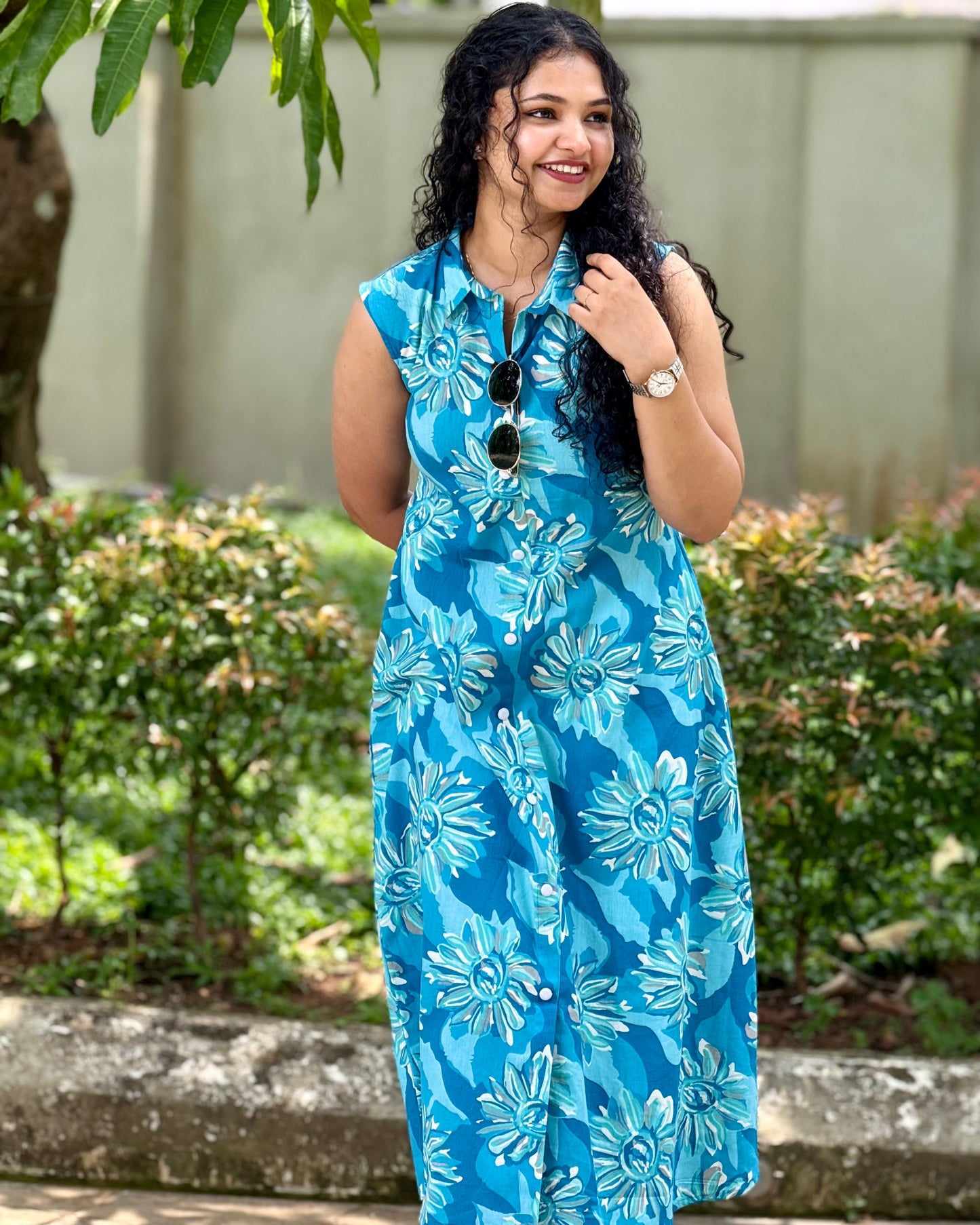 Woman wearing a blue floral dress standing outdoors with greenery in the background