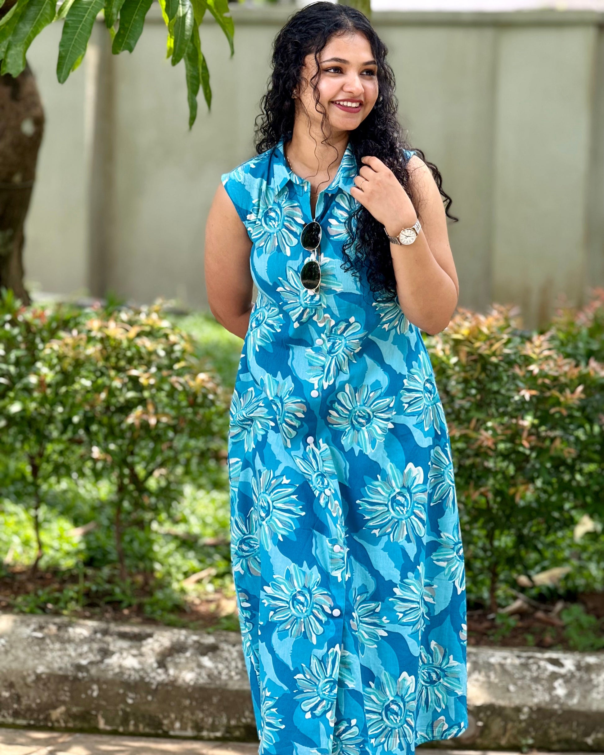 Woman wearing a blue floral dress standing outdoors with greenery in the background