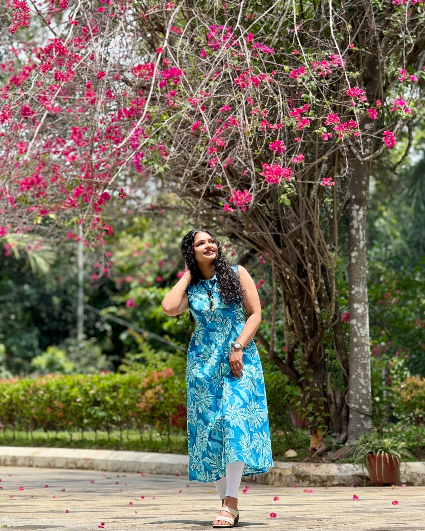 Woman in a blue dress standing under a pink flowering tree in a park.