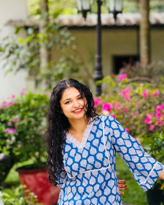 Woman in a blue patterned dress standing in a garden with pink flowers and a blurred building in the background.