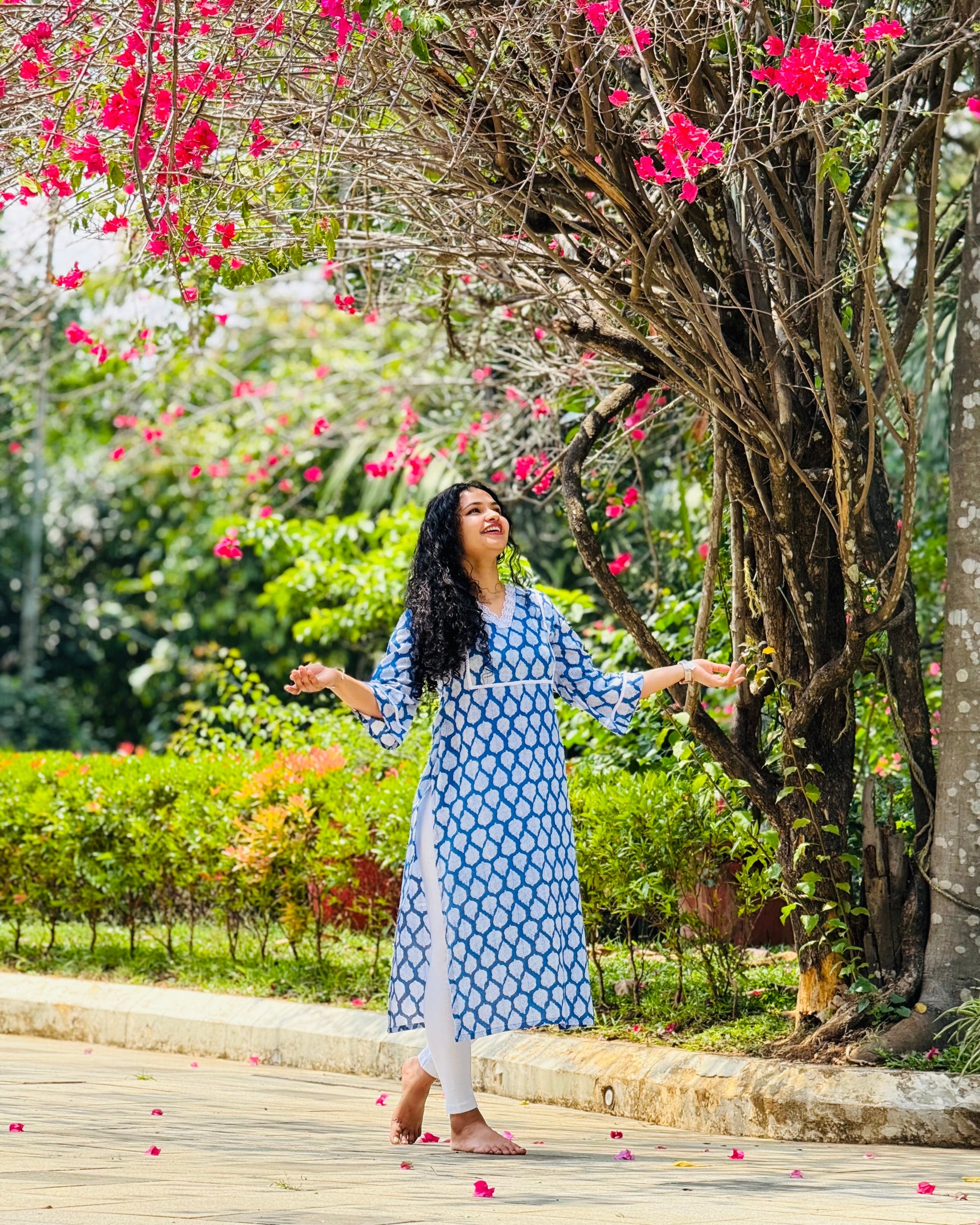 Woman in a blue dress standing under a pink flowering tree in a garden.