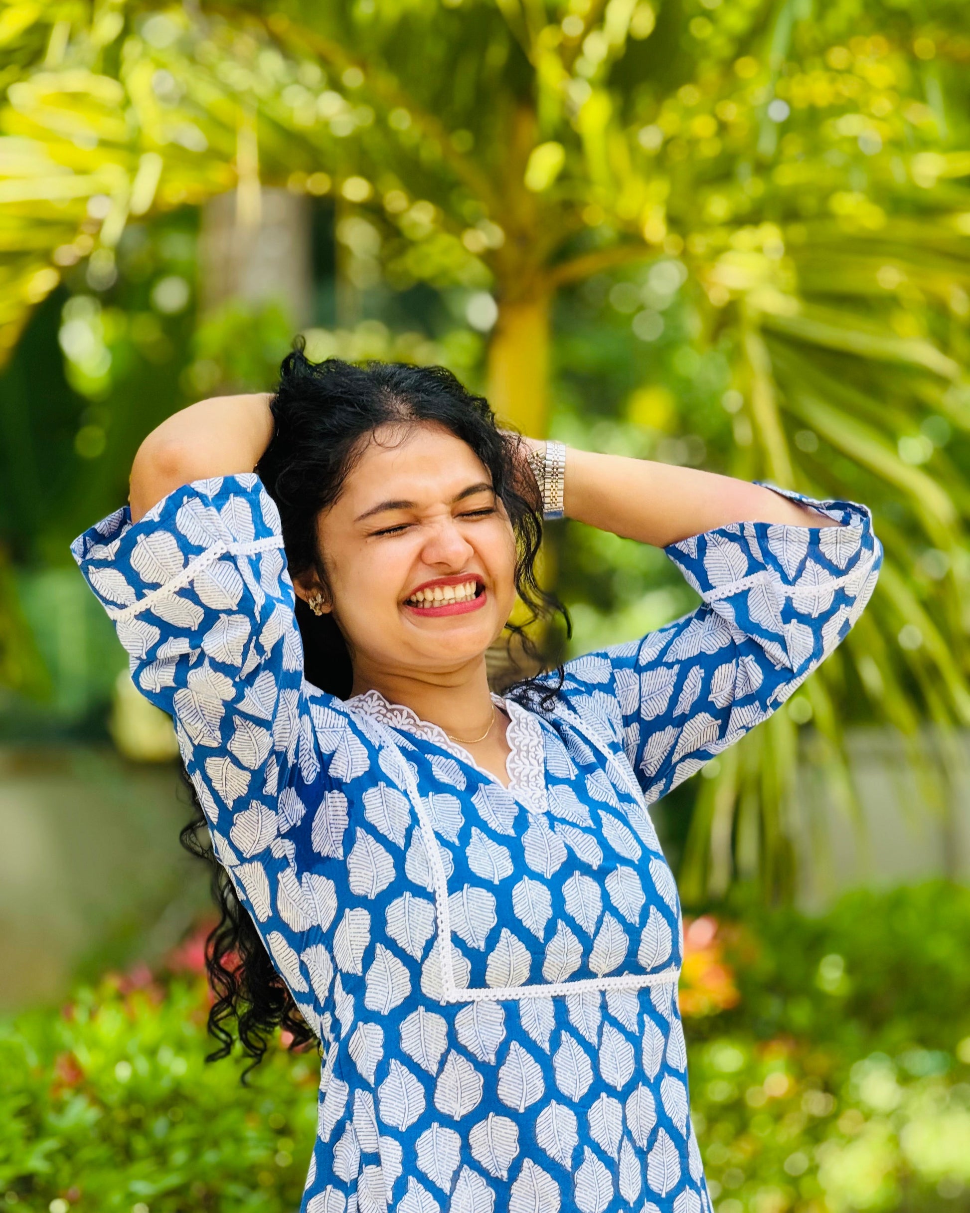 Woman in a blue and white patterned dress standing outdoors with greenery in the background