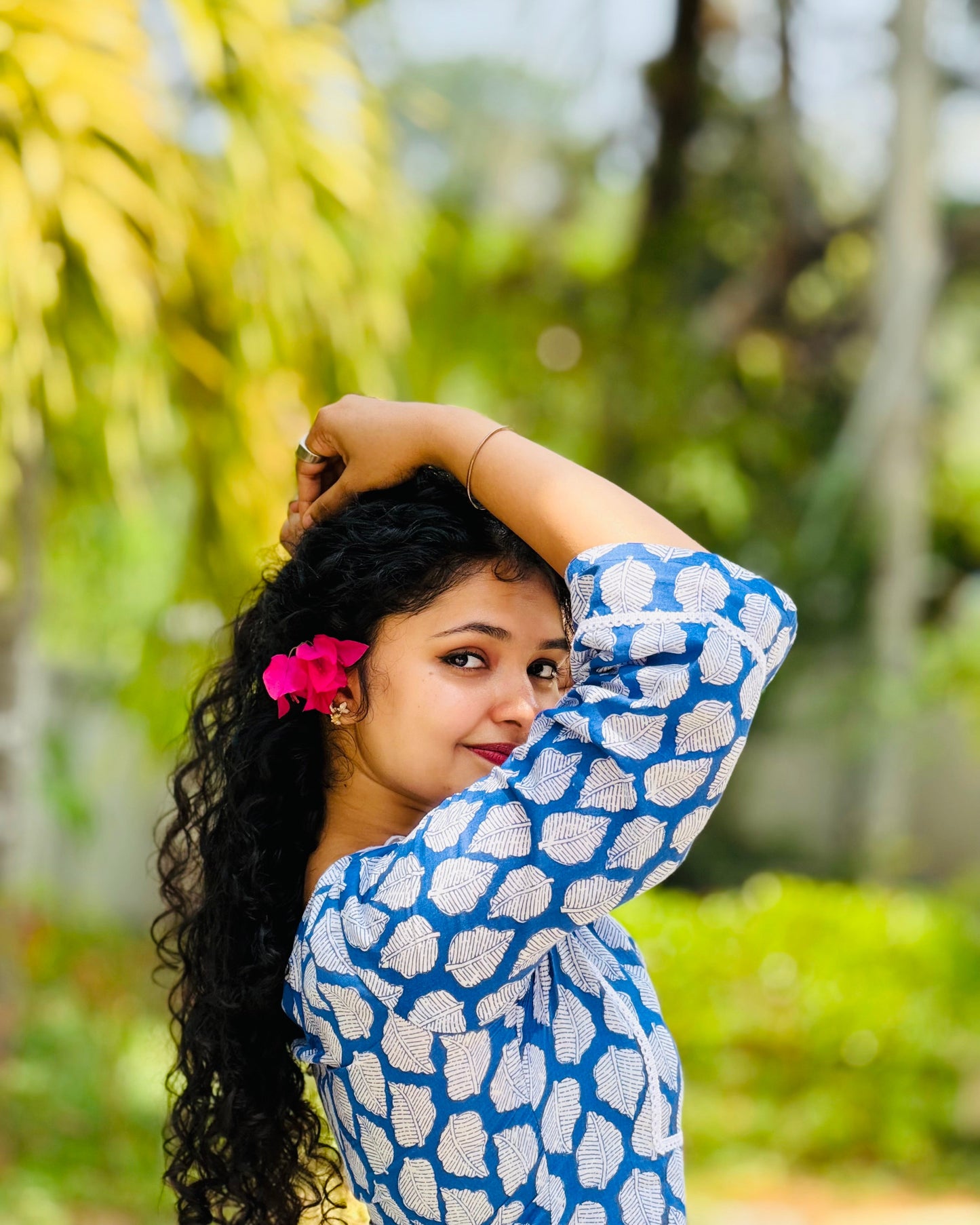 Woman in a blue and white patterned top with a pink flower in her hair, standing outdoors with greenery in the background.