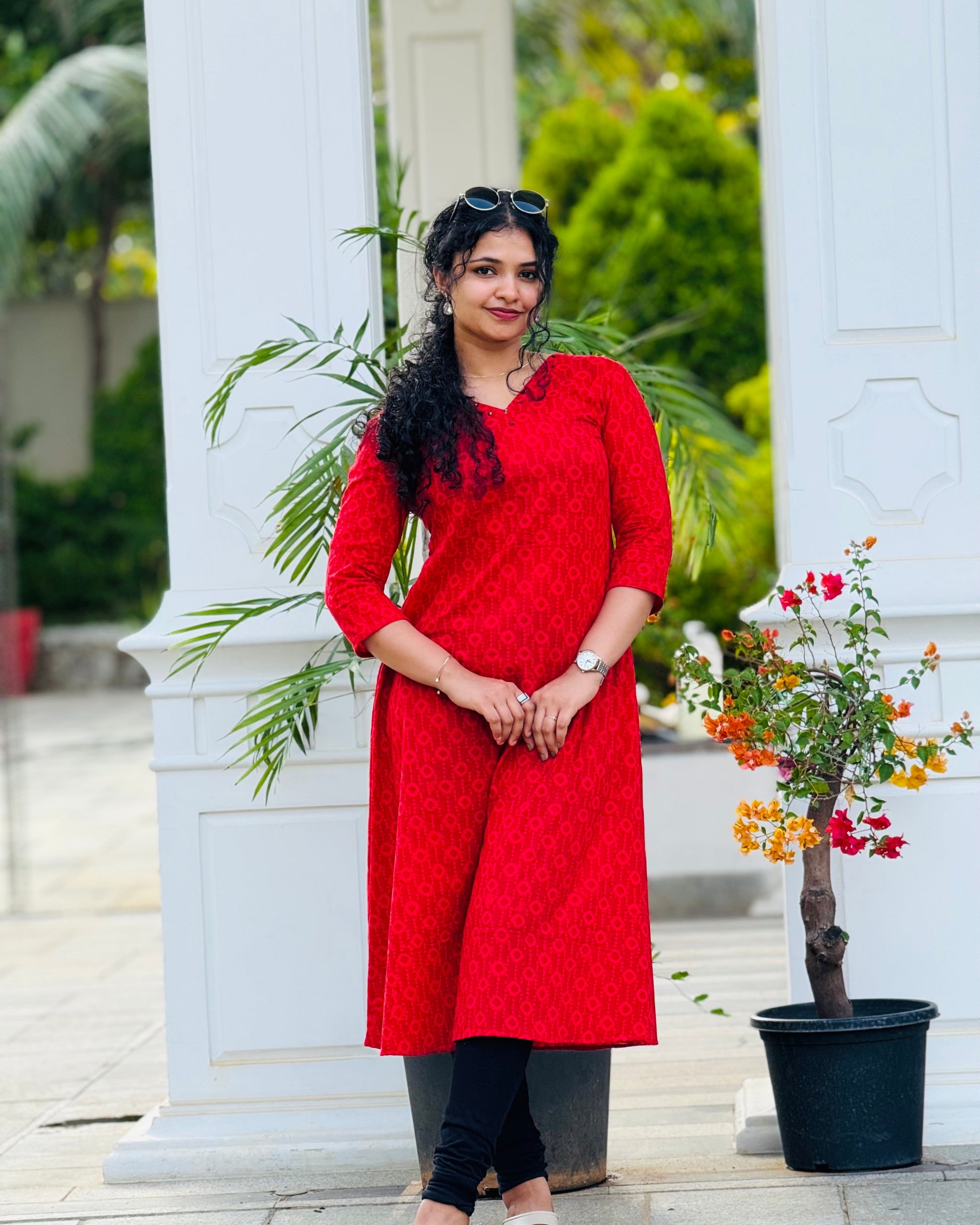 Woman in a red kurta standing outdoors with greenery and a white architectural structure in the background.