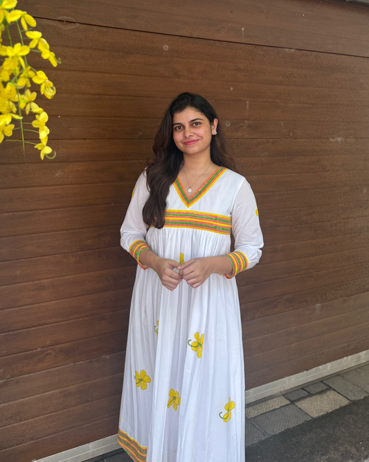 Woman in a white dress with yellow patterns standing against a wooden wall with yellow flowers.