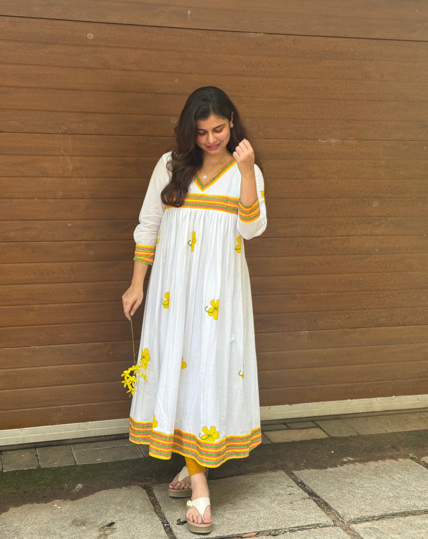 Woman wearing a white dress with yellow patterns against a wooden wall.