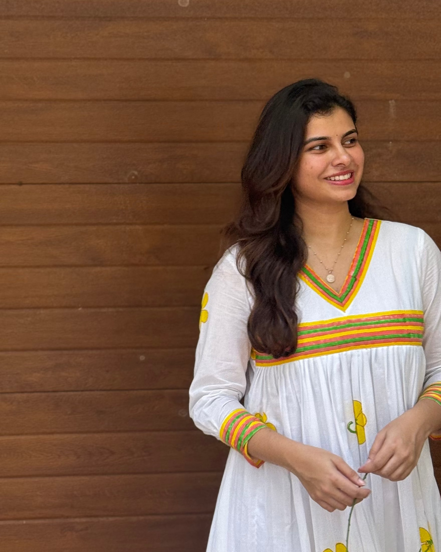 Woman in a white traditional outfit with colorful accents standing against a wooden wall.