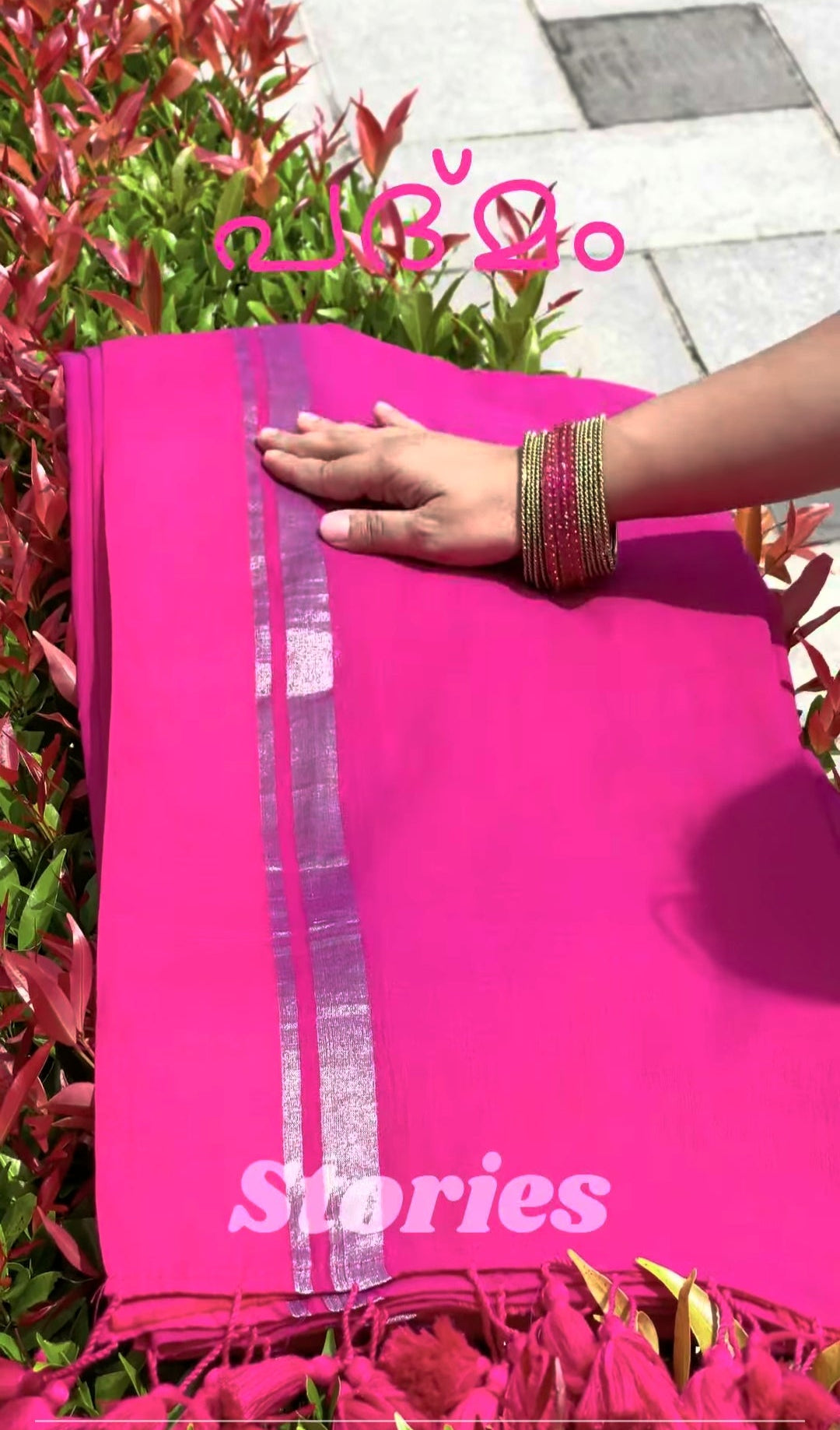 Pink saree with 'Stories' branding held by a hand against a green plant background