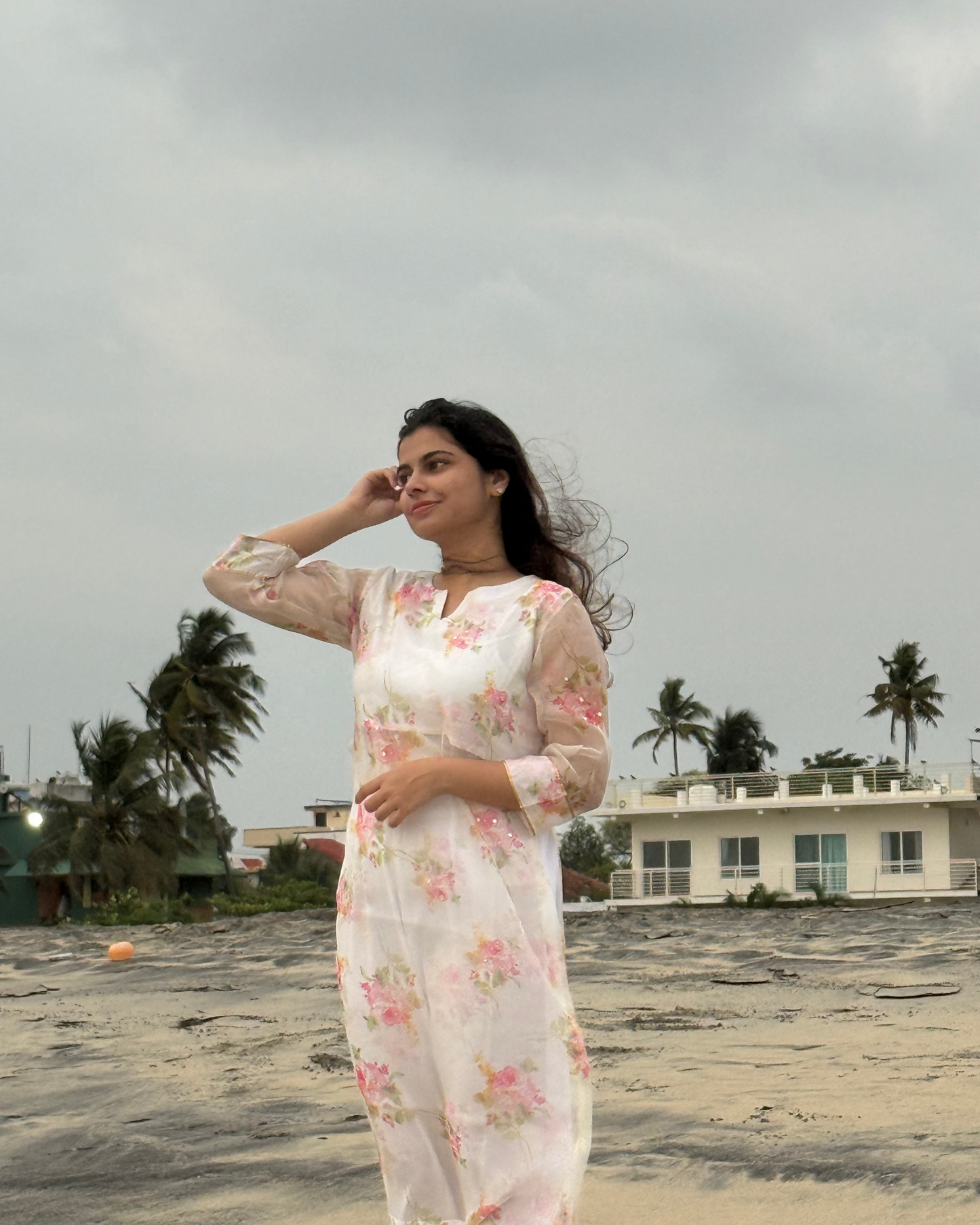Woman in a floral dress standing on a beach with palm trees and a building in the background.