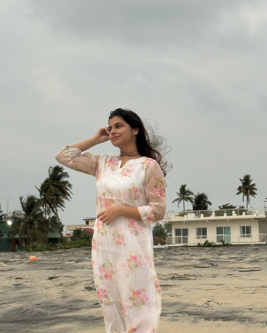 Woman in a floral dress standing on a beach with palm trees and a building in the background.